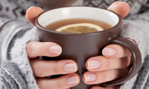 O persoană, îmbrăcată într-un pulover de iarnă tricotat, ține cu ambele mâini o cană maro de ceai cald, din care aburii se ridică ușor. O felie subțire de lămâie plutește în băutură, sugerând un remediu reconfortant pentru răceală sau o băutură caldă, perfectă pentru serile friguroase de iarnă. Mâinile sunt îngrijite, iar nuanțele calde ale cănii contrastează plăcut cu textura moale a puloverului.