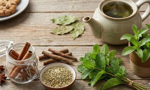 Remedii naturale pe o masă de lemn, incluzând scorțișoară, fenicul, mentă, lemn dulce, un ghiveci cu stevia și ceai verde, alături de o farfurie cu prăjituri, ilustrând plante care reduc pofta de dulciuri.