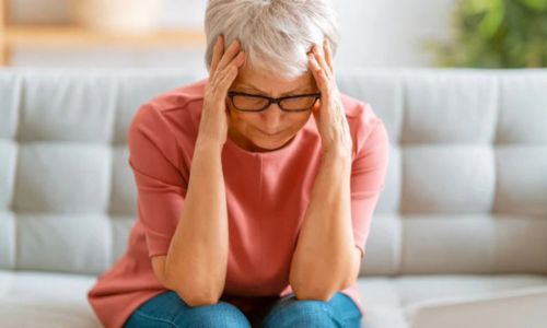  O femeie în vârstă, cu părul alb și ochelari, stă pe o canapea cu o expresie de disconfort, ținându-se de cap cu ambele mâini. Pare să sufere de o durere de cap.