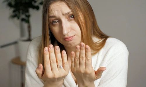 Imaginea surprinde o femeie tânără, îmbrăcată în halat alb, care își privește îngrijorată vârfurile degetelor. Expresia feței indică nemulțumire sau îngrijorare, posibil legată de starea unghiilor sau a pielii de pe degete. Fundalul este simplu, cu o plantă verde în colț, sugerând un cadru casnic. Această imagine poate ilustra îngrijorări legate de sănătatea unghiilor, pielea uscată sau probleme dermatologice la mâini.